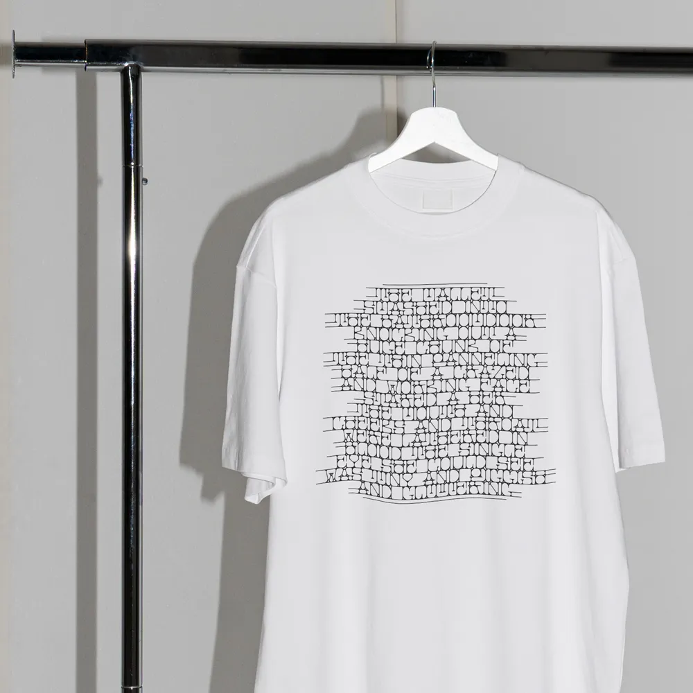 White t-shirt hanging on a white hanger from a metal rack, with abstract black text printed in a grid pattern on the front.